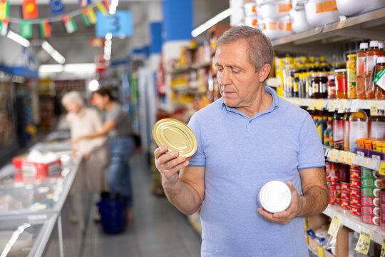 Aging man in blue t-shirt intently focused on selecting right canned food product, carefully examining labels on tin cans. Concept of shopping experience and informed decision making..