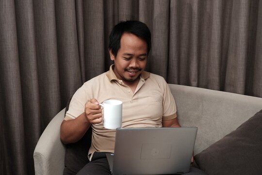 Southeast Asian man in a beige shirt sitting on a sofa, smiling while using a laptop and holding a white coffee mug in a relaxed setting.