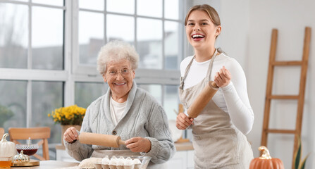 Young woman and her grandmother with rolling pins preparing dough in kitchen
