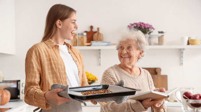 Young woman holding baking tray with cookies and her grandmother in kitchen - Powered by Adobe