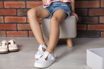 Little girl trying on different shoes near brick wall indoors, closeup