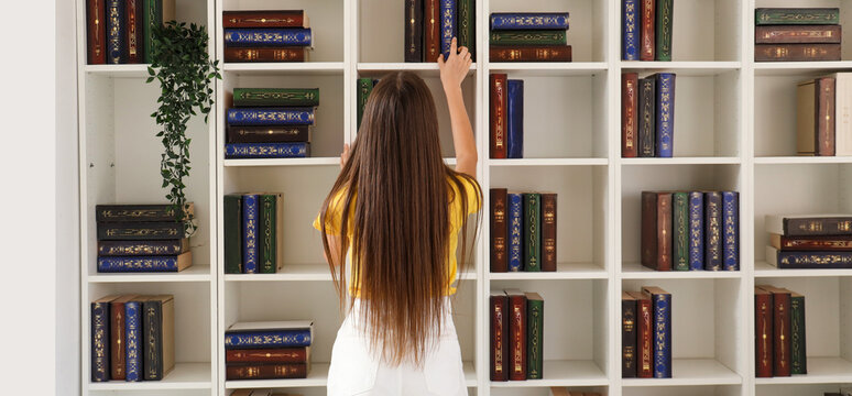 Female student taking book from shelving unit while studying at library, back view