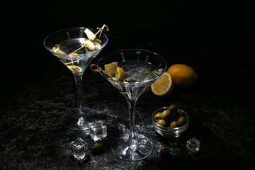 Glasses of tasty martini, ice cubes, green olives and lemon on dark background