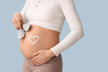 Young pregnant woman with heart made of cream on her belly and baby booties against blue...