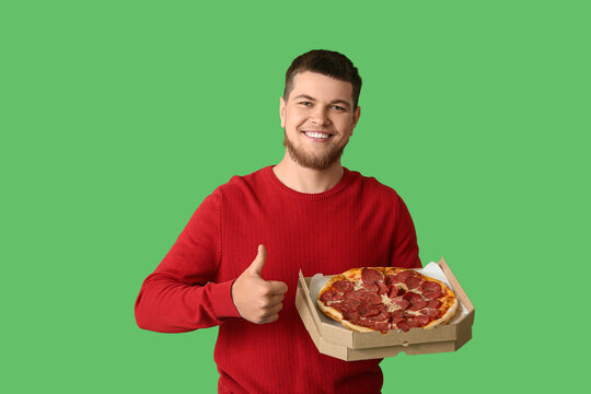 Young man holding cardboard box with tasty pepperoni pizza showing thumb-up gesture on green background
