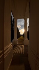 Sunburst shining over a bright green park alley, framed by the dark silhouette of a classical portico's columns and railings, emphasizing contrast