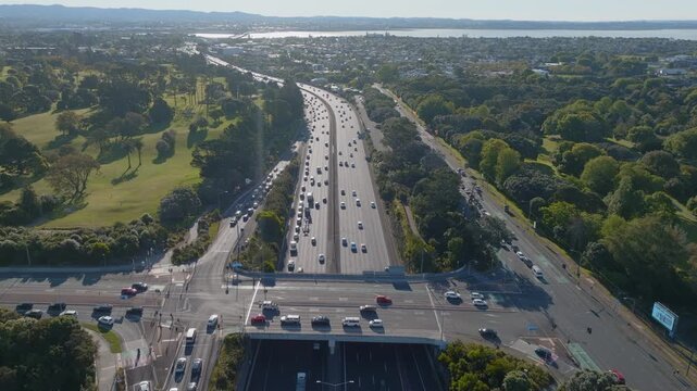 Aerial view of cars traveling on a busy highway in Auckland, New Zealand. The highway is surrounded by lush green trees and a golf course, with a city skyline in the distance.