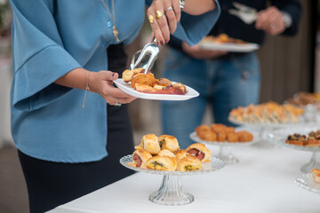 A woman in a blue blouse serves herself mini sausage rolls from a crystal cake stand at an elegant catered event with assorted appetizers displayed on a white tablecloth in soft natural light