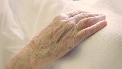 The wrinkled hand of an old woman lying in bed is gently held by the hand of a man. Themes of aging, death, caring, love, family. 