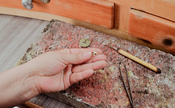 Jeweler artisan hand holding small metal jewelry piece on workbench with tools and firebrick, handmade jewelry process, traditional metalworking and precision craftsmanship. - Powered by Adobe