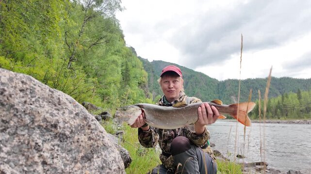 Female fisher celebrates a successful catch in the mountains &mdash; close-up of the taimen&rsquo;s vivid tail and open mouth, with forested slopes behind, until it slips free, drops to the ground