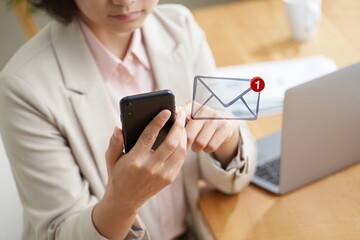 Asian woman in a light blazer is focused on her smartphone, checking email notifications while seated at a wooden desk with a laptop and office supplies, creating a professional atmosphere