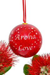 A red Christmas bauble seen against a white background framed by the striking red flowers of New Zealand's Summer flowering Pohutukawa tree. The tree is known as the NZ Christmas tree.