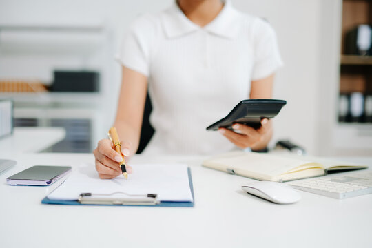 A young office woman calculating finances with a calculator and notebook at her desk, representing accounting, budgeting, financial planning, and office productivity. - Powered by Adobe