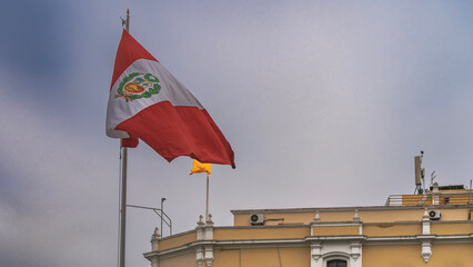 The national flag of Peru is flying against the sky. A red and white banner with the coat of arms of the country. A fragment of a building Municipal Palace of Lima