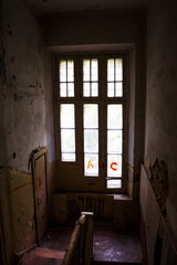 Eerie abandoned room showcasing a large, dirty window with red graffiti. Peeling paint, decaying walls, and a dark staircase suggest neglect and mystery
