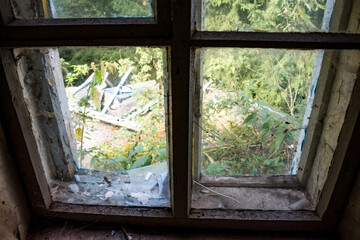 Fototapeta premium Decaying window frame with shattered glass and peeling paint, offering a gritty glimpse of lush, untamed nature reclaiming an abandoned space