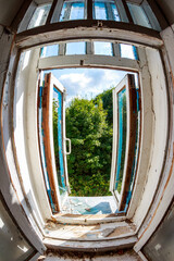 Dilapidated wooden window frame inside abandoned building, wide-angle view. Open panes reveal vibrant summer greenery and sky. Broken glass, peeling paint, urban decay