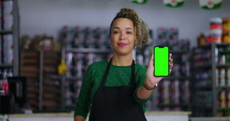 Shopkeeper holding a smartphone with a green screen and chroma key in a hardware store, presenting the device towards the camera, shelves of colorful products in the background