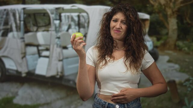 Woman holding green apple and clutching stomach in forest near parked cart at dusk; discomfort hesitation.