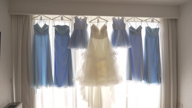 A stunning display of blue and white dresses is hung in front of a bright window, highlighting their delicate fabric and elegant designs. Perfect for a special occasion.
