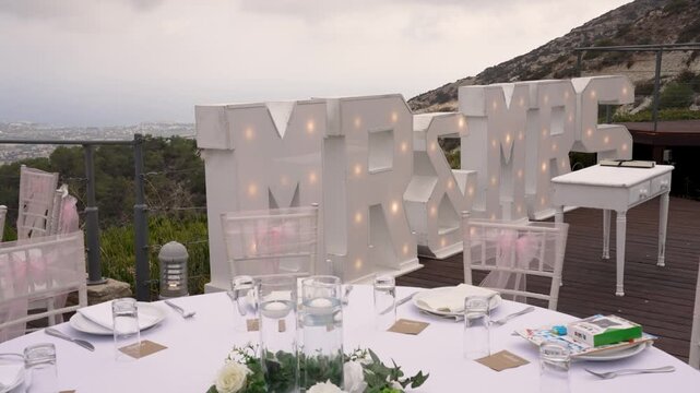 A stunning wedding reception is arranged outdoors with a breathtaking mountain view. The table is set with white decorations, flowers, and illuminated letters declaring Mr and Mrs.
