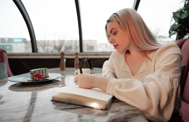 Dreamy young woman writing down future plans and goals in own diary resting in cozy cafeteria