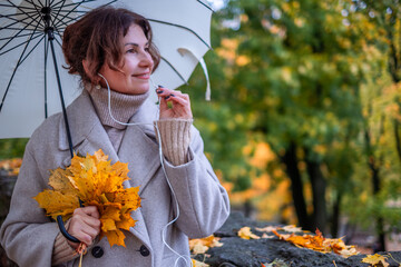 Midlife woman with curly hair holding yellow autumn leaves and an umbrella, enjoying a call outdoors in a vibrant fall setting with colorful foliage
