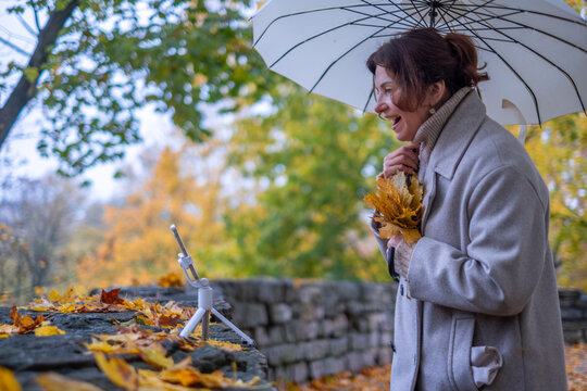 Midlife woman holding autumn leaves under a white umbrella, smiling while taking a selfie with a smartphone on a tripod in a colorful fall landscape with vibrant foliage - Powered by Adobe