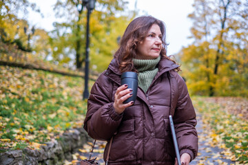 Middle-aged woman in brown jacket holding coffee cup and laptop, walking through autumn park with colorful leaves, embodying the concept of working while enjoying nature
