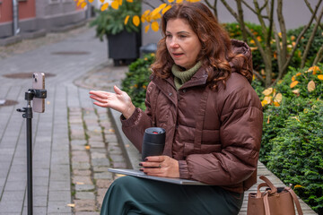 Middle-aged woman engaged in video call meeting outdoors, wearing a brown puffer jacket, holding a coffee cup, with greenery and autumn leaves in the background, showcasing remote work concept