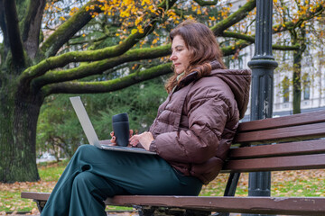Midlife woman engaged in video call meeting while working remotely on laptop in a park, surrounded by autumn foliage and natural scenery, showcasing modern work-life balance