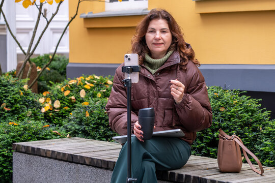 Middle-aged woman engaged in video call meeting outdoors, seated on a bench with greenery around, showcasing modern business communication and remote work concept