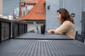 Middle-aged woman working remotely at a food court table, focused on her laptop, with a coffee cup beside her, showcasing a modern work environment and lifestyle