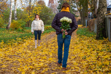 Midlife couple walking through autumn park, man holding bouquet of roses behind back, woman smiling, surrounded by colorful leaves, capturing romantic moment in nature