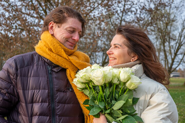 Midlife couple sharing a joyful moment outdoors, surrounded by autumn foliage, holding a bouquet of white roses, celebrating love and connection in a serene setting with copy space