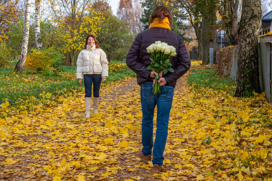 Midlife couple walking through autumn park, man holding bouquet of roses behind back, woman smiling, surrounded by colorful leaves, capturing romantic moment in nature
