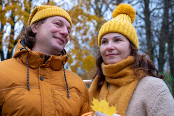 Middle-aged couple in cozy autumn attire smiles while holding colorful leaves, surrounded by vibrant fall foliage, enjoying a peaceful moment in nature during the autumn season