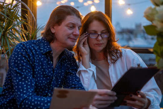 Middle-aged couple enjoying a cozy evening in a cafe, reviewing a menu together, surrounded by warm lighting and a relaxed atmosphere, capturing a romantic moment - Powered by Adobe