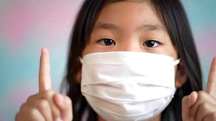 Closeup of a young Asian girl wearing a white protective face mask looking directly at the camera and pointing upwards with both index fingers symbolizing protection and awareness during a pandemic.