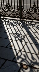 The intricate shadow of an ornate wrought iron gate cast onto tiled ground creates a striking pattern with natural light and shadows