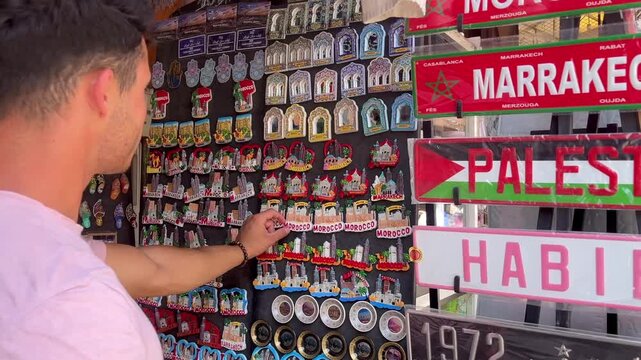 Latin european young male tourist shopping at an outdoor market stall in Marrakech, Morocco, choosing a fridge magnet as a keepsake of his memorable vacation and travel experience in Africa