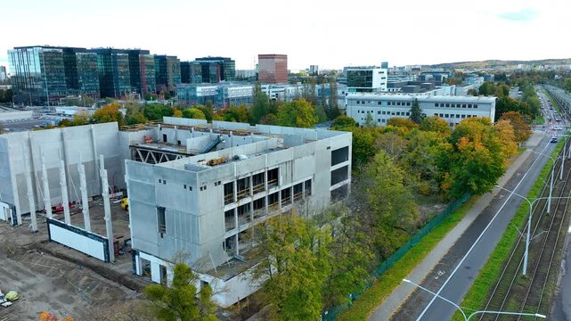 large construction site surrounded by trees and modern glass office buildings in Gdańsk, Poland. The site shows the emerging concrete structure of a sports or research facility under development
