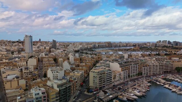 Orbital drone shot circling above Msida and Ta&rsquo; Xbiex, Malta, revealing Gżira and Sliema skylines with marinas, yachts, and developing coastal architecture under a blue cloudy sky.