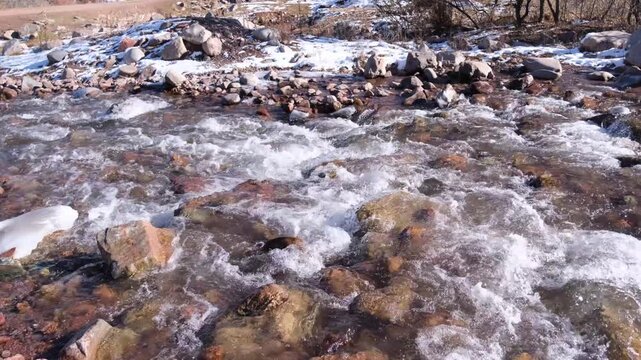 Clear mountain river flowing over brown rocks and pebbles, with small patches of snow on the bank. Winter nature scenery.