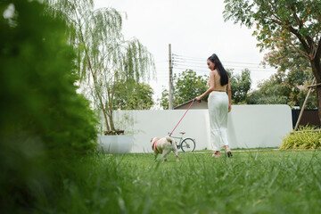 Asian woman walking her pug dog on leash in green garden, enjoying outdoor activity and relaxing lifestyle with nature.