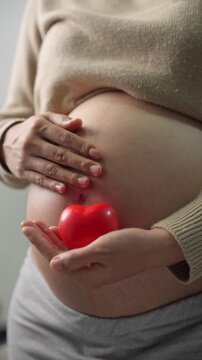 Happy pregnant woman or mother touching baby in belly and hold red heart with love at home. Health care, Prenatal preparation, Maternity, Pregnancy, and motherhood concept
