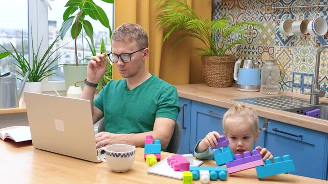 Father works remotely in the kitchen on a laptop, daughter little child plays with a construction kit nearby, without interfering with my dad's work.