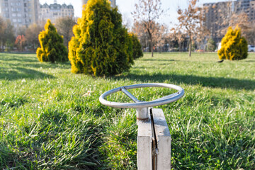 A close-up view of a metal valve wheel is shown in a grassy urban park. Evergreen shrubs and tall buildings are visible in the background under clear skies.