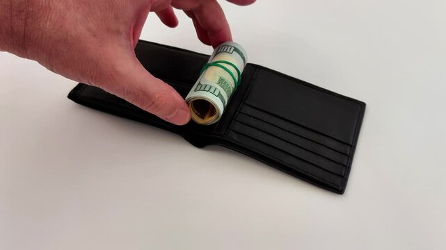 Hands tuck rolled-up 100 bills into a sleek black wallet resting on a white table-clean setup, crisp motion, money-focused action with minimalist contrast